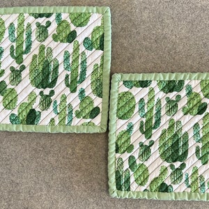 May include: Two square quilted pot holders featuring a white background with a repeating pattern of green cacti. The edges are trimmed with a light green fabric. Diagonal stitching adds texture to the pot holders.
