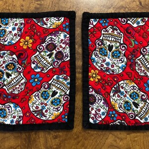 May include: Two red quilted pot holders with a black border. The pot holders feature a repeating pattern of white sugar skulls with colourful floral designs.