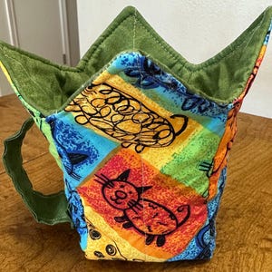 May include: A colorful fabric mug cozy with a green handle and a green, pointed top. The cozy features a vibrant pattern of cats and abstract shapes in orange, blue, yellow, and green. The cozy is designed to insulate a mug.