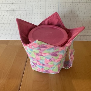 Op de afbeelding: Een roze en paarse bloemenstofcontainer met een handvat. De container bevat een roze plastic container met een deksel.