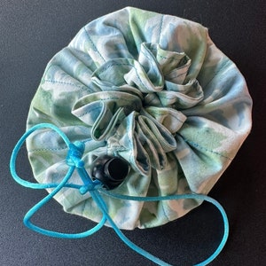 May include: A round, fabric drawstring bag with a floral pattern in shades of blue and green. The bag has a gathered top and a light blue drawstring with a black toggle closure.