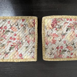 May include: Two beige quilted pot holders with a fall leaf and acorn print. The pot holders have a tan border.