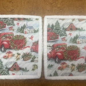 May include: A pair of red and white quilted pot holders with a winter holiday theme. The pot holders feature red trucks with Christmas trees and presents, along with snow-covered houses and trees.