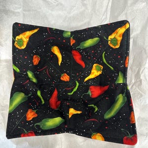 Puede incluir: Un protector de cuenco de tela negra con un diseño acolchado. El protector tiene un estampado de pimientos picantes de colores en tonos rojos, amarillos, naranjas y verdes. Hay motas blancas dispersas en el fondo.