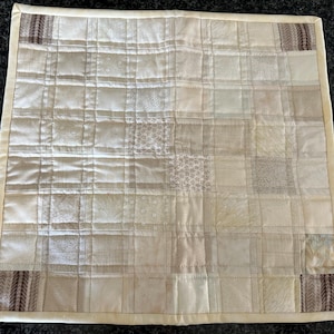 Può includere: Un sottopentola quadrato e trapuntato con un design patchwork. Presenta una varietà di tessuti bianco sporco e beige, con un bordo color crema. Gli angoli hanno un tessuto a righe marrone e beige. Le cuciture sono visibili.