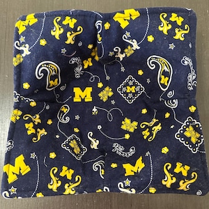 Op de afbeelding: Marineblauwe pannenlap met een herhalend patroon van gele "M" logo's, paisley vormen en bloemmotieven. De pannenlap is vierkant met een zak aan één kant. Het ontwerp is gedrukt op een donkerblauwe stof.