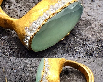 Puede incluir: Un anillo de oro con una gran piedra preciosa ovalada de color verde claro engastada en el centro. La piedra preciosa está rodeada por una fila de pequeños diamantes de imitación transparentes.