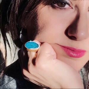 Puede incluir: Un anillo de oro con una gran piedra ovalada turquesa engastada en un engaste de plata. La piedra est&aacute; rodeada por una fila de peque&ntilde;os diamantes.
