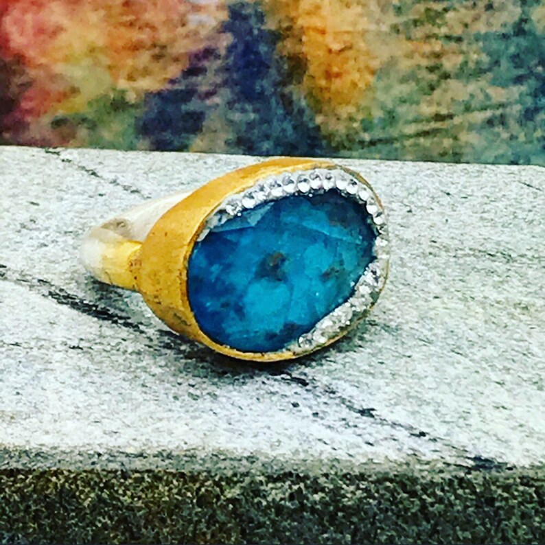 Puede incluir: Un anillo de oro con una gran piedra ovalada turquesa engastada en un engaste de bisel. La piedra est&aacute; rodeada por una fila de peque&ntilde;os diamantes blancos.