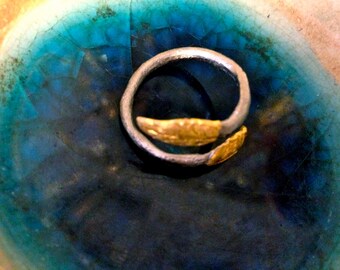 Puede incluir: Un anillo plateado con dos detalles de hojas doradas. El anillo se encuentra sobre un fondo de tonos azules y turquesas, creando un contraste llamativo. El diseño del anillo es simple pero elegante, adecuado para el uso diario.
