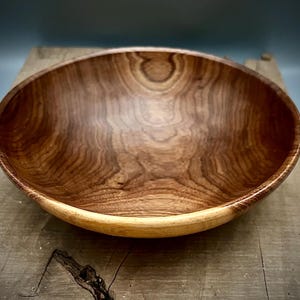Puede incluir: Un cuenco de madera hecho a mano con un patrón de vetas de madera natural. El cuenco tiene forma ovalada con un borde de color claro y un interior marrón intenso. El cuenco está sobre una superficie de madera.