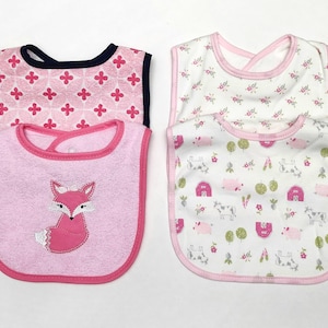 May include: Four baby bibs in the image. Two bibs have pink and white floral patterns, and two have pink backgrounds. One pink bib has a fox applique, and the other has a pink floral pattern with a navy blue trim.