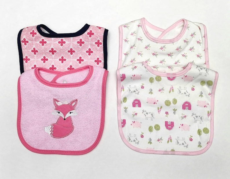 May include: Four baby bibs in the image. Two bibs have pink and white floral patterns, and two have pink backgrounds. One pink bib has a fox applique, and the other has a pink floral pattern with a navy blue trim.