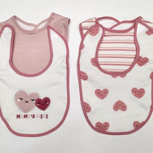 May include: Two baby bibs. One bib is white with pink trim and features two hearts and the text "Mama's Mini". The other bib is white with pink hearts and pink striped accents.