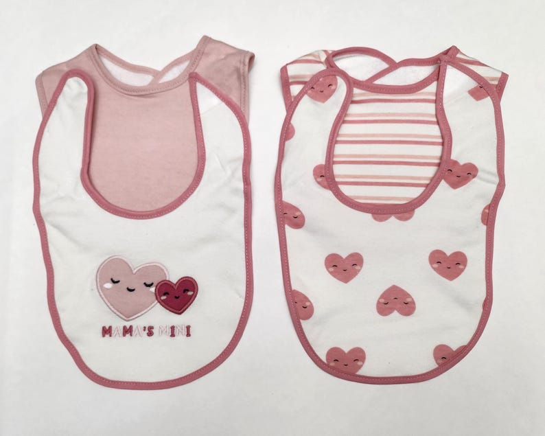 May include: Two baby bibs. One bib is white with pink trim and features two hearts and the text "Mama's Mini". The other bib is white with pink hearts and pink striped accents.