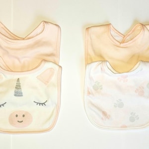 May include: Two baby bibs. One is white with a unicorn face design, including a silver horn, pink ears, and closed eyes. The other is white with a pattern of pink and gray paw prints. Both have a peach-colored backing.