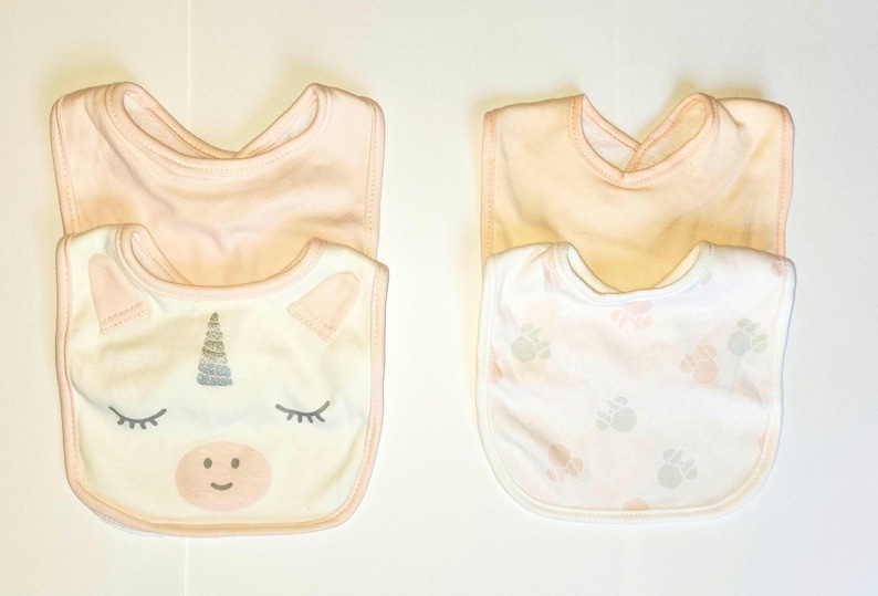 May include: Two baby bibs. One is white with a unicorn face design, including a silver horn, pink ears, and closed eyes. The other is white with a pattern of pink and gray paw prints. Both have a peach-colored backing.