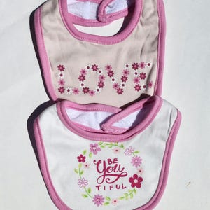 May include: Two baby bibs. The top bib is tan with pink trim and the word "LOVE" spelled out in pink floral letters. The bottom bib is white with pink trim and the words "Be You Tiful" surrounded by pink flowers.