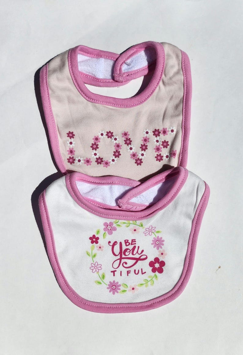 May include: Two baby bibs. The top bib is tan with pink trim and the word "LOVE" spelled out in pink floral letters. The bottom bib is white with pink trim and the words "Be You Tiful" surrounded by pink flowers.