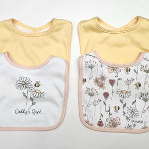 May include: Two baby bibs with a yellow top and a white bottom. One bib features a daisy and bee design with the text "Daddy's Girl". The other bib has a floral pattern with bees. Both have pink trim.