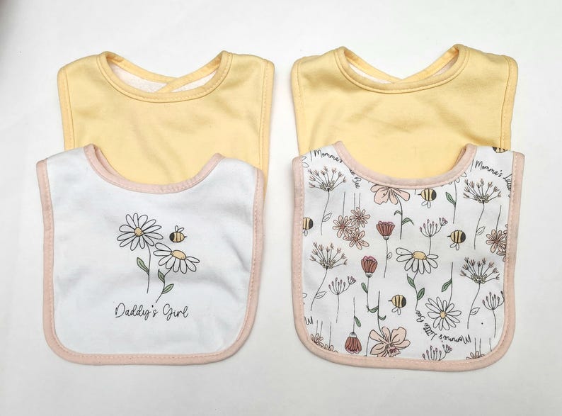 May include: Two baby bibs with a yellow top and a white bottom. One bib features a daisy and bee design with the text "Daddy's Girl". The other bib has a floral pattern with bees. Both have pink trim.