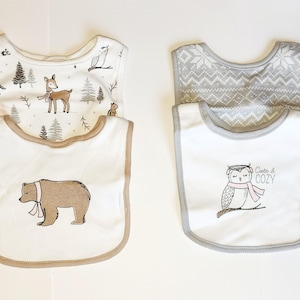 May include: Four baby bibs with woodland animal designs. Two bibs have a white background with a bear and owl graphic. The other two bibs have a forest and snowflake pattern. The owl bib says "Cute & COZY".