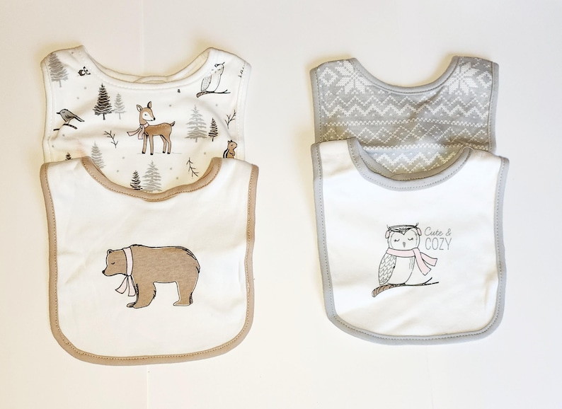May include: Four baby bibs with woodland animal designs. Two bibs have a white background with a bear and owl graphic. The other two bibs have a forest and snowflake pattern. The owl bib says "Cute & COZY".