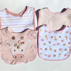 May include: Four baby bibs in soft pastel colors. One bib has a giraffe face with a pink bow. Another has a white background with pink and yellow stripes. The other two bibs have animal patterns and pink trim.