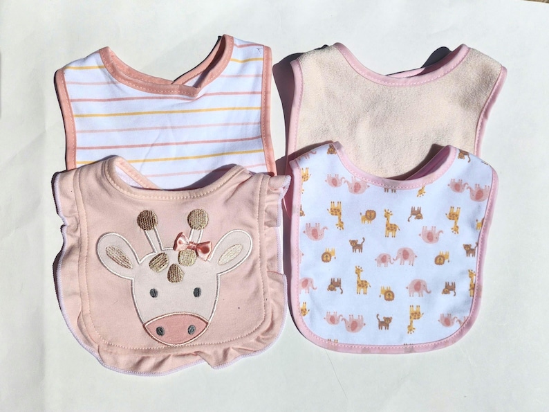 May include: Four baby bibs in soft pastel colors. One bib has a giraffe face with a pink bow. Another has a white background with pink and yellow stripes. The other two bibs have animal patterns and pink trim.