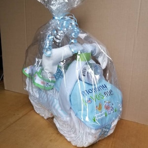 May include: A baby gift basket shaped like a motorcycle, wrapped in clear cellophane and tied with a blue ribbon. The basket includes diapers, a bottle, and a blue bib with the text "Mommy loves me" and farm animal graphics.