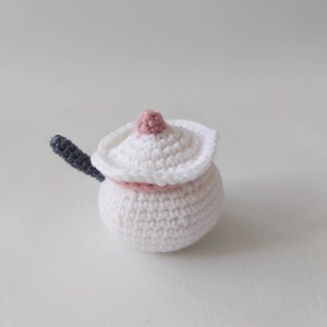 May include: A white crocheted sugar bowl with a pink lid and a gray handle. The bowl is round and has a textured surface.