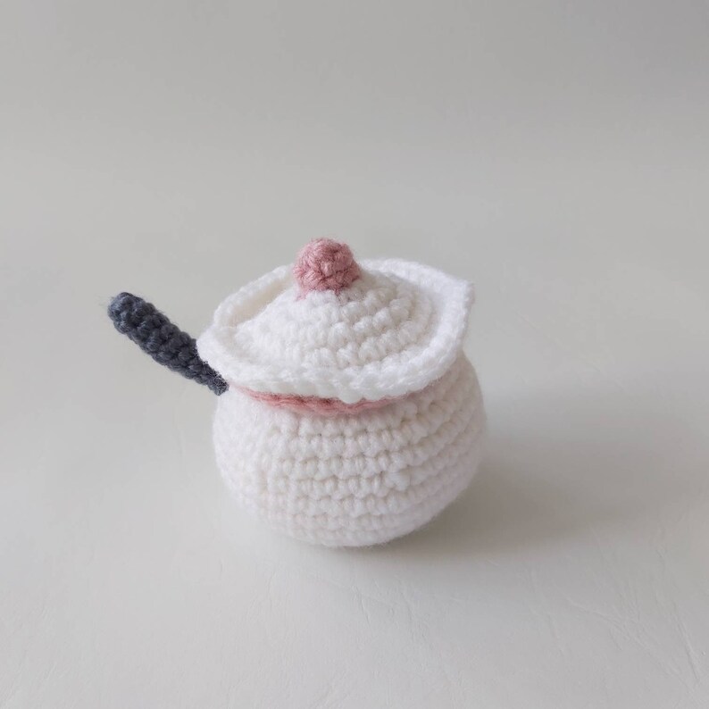 May include: A white crocheted sugar bowl with a pink lid and a gray handle. The bowl is round and has a textured surface.