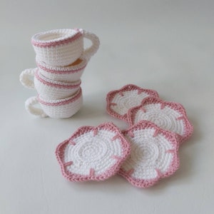 May include: Set of four crocheted coasters in white with a pink trim. The coasters are shaped like a flower with five petals. There are also four crocheted teacups stacked on top of each other, also in white with a pink trim.