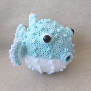 Puede incluir: Un juguete de pez globo de crochet azul y blanco con ojos negros.