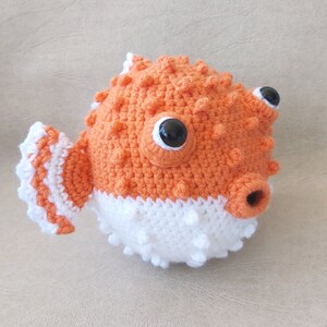 Puede incluir: Un juguete de pez globo de crochet naranja y blanco con ojos negros. El juguete tiene una superficie texturizada y una peque&ntilde;a boca.