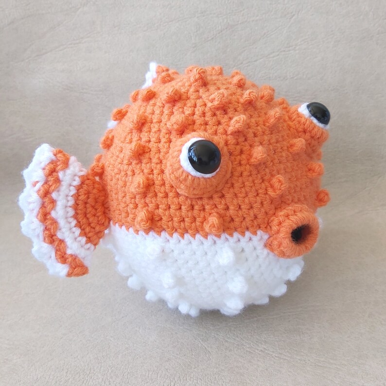 Puede incluir: Un juguete de pez globo de crochet naranja y blanco con ojos negros. El juguete tiene una superficie texturizada y una peque&ntilde;a boca.