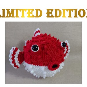 Puede incluir: Un juguete de peluche de pez globo rojo y blanco tejido a crochet con ojos negros. El juguete es una edici&oacute;n limitada.