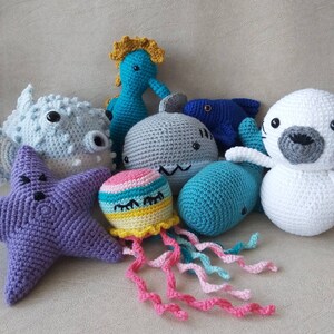 Puede incluir: Una colecci&oacute;n de criaturas marinas de crochet, que incluyen una estrella de mar p&uacute;rpura, un tibur&oacute;n gris, un caballito de mar azul, un pez azul, una foca blanca, una ballena turquesa y una medusa arco&iacute;ris.