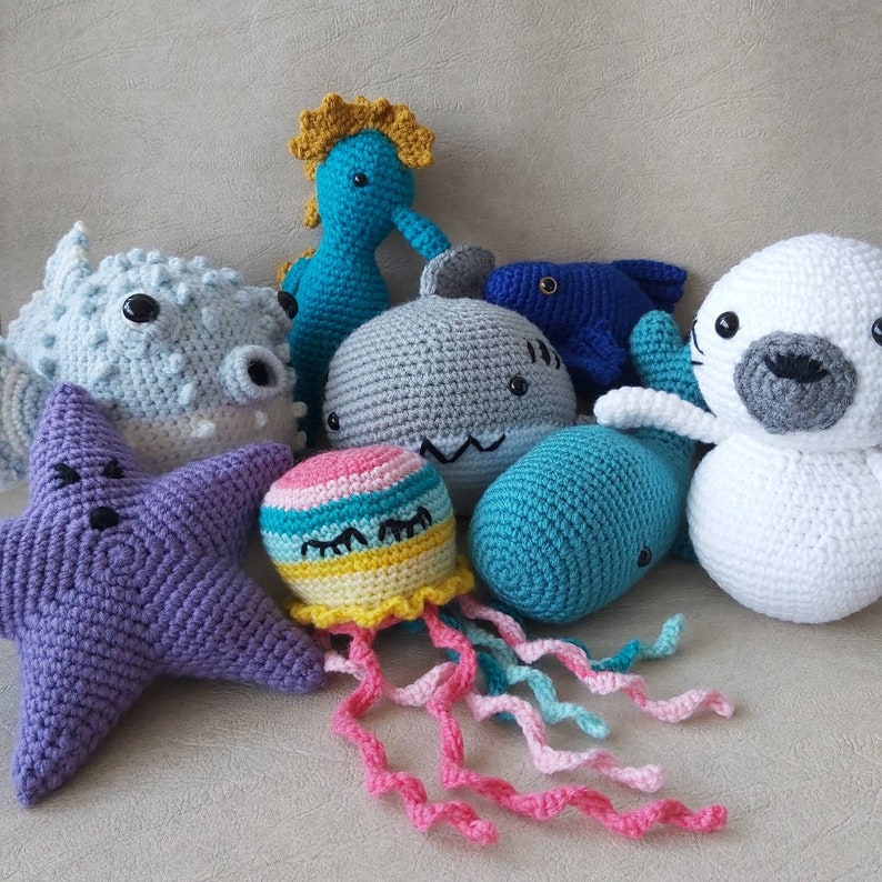 Puede incluir: Una colecci&oacute;n de criaturas marinas de crochet, que incluyen una estrella de mar p&uacute;rpura, un tibur&oacute;n gris, un caballito de mar azul, un pez azul, una foca blanca, una ballena turquesa y una medusa arco&iacute;ris.