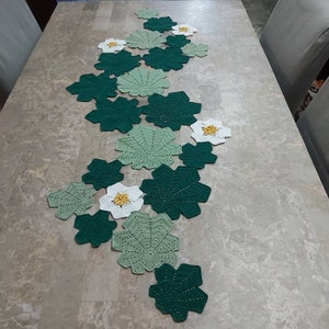 May include: A crocheted table runner with green and white leaves and flowers. The runner is made of a delicate, lacy yarn and is perfect for adding a touch of elegance to any table.