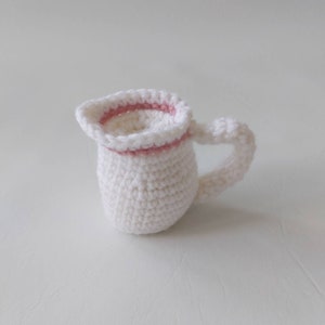 May include: A white crocheted pitcher with a pink crocheted rim. The pitcher has a handle and is a small size.
