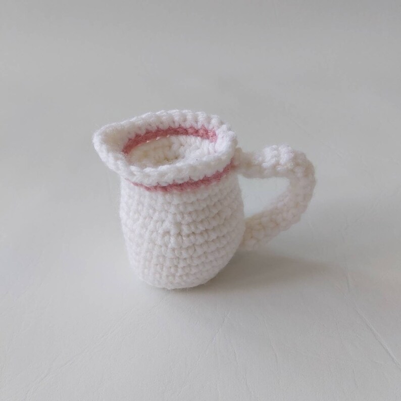 May include: A white crocheted pitcher with a pink crocheted rim. The pitcher has a handle and is a small size.