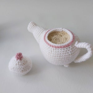 May include: A white crocheted teapot with a pink crocheted rim and a matching white crocheted lid with a pink crocheted top.