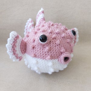 Puede incluir: Un juguete de pez globo de crochet rosa y blanco con ojos negros.
