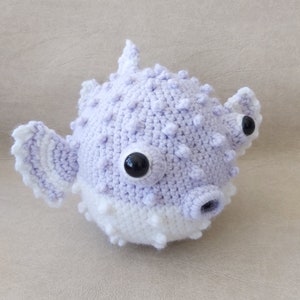 Puede incluir: Un juguete de peluche de pez globo de color morado y blanco tejido a crochet con ojos negros.