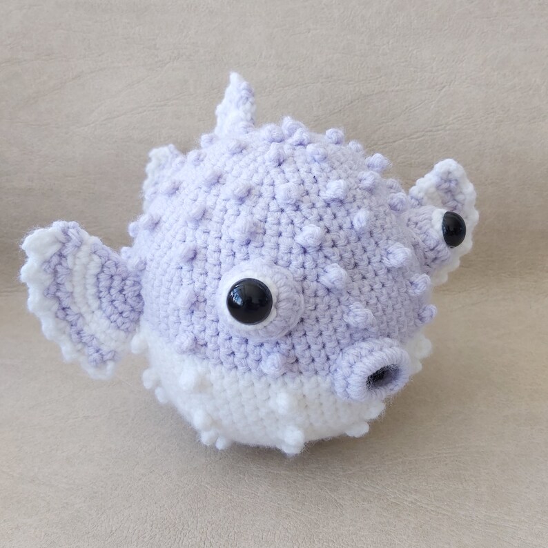 Puede incluir: Un juguete de peluche de pez globo de color morado y blanco tejido a crochet con ojos negros.
