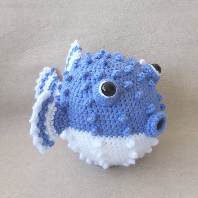 Puede incluir: Un juguete de pez globo de crochet azul y blanco con ojos negros. El pez globo tiene la barriga blanca y las aletas azules.