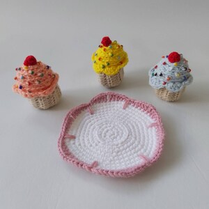 May include: Three crocheted cupcakes in pink, yellow, and blue with colorful sprinkles and red cherries on top. The cupcakes are sitting on a white crocheted plate with a pink border.