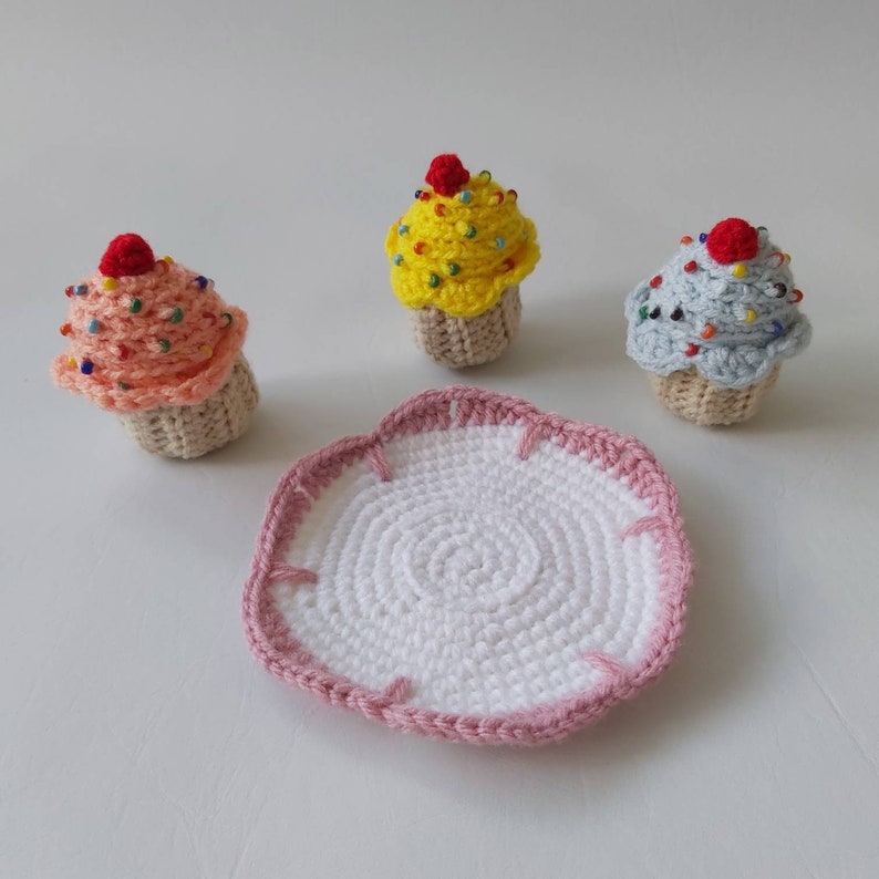May include: Three crocheted cupcakes in pink, yellow, and blue with colorful sprinkles and red cherries on top. The cupcakes are sitting on a white crocheted plate with a pink border.