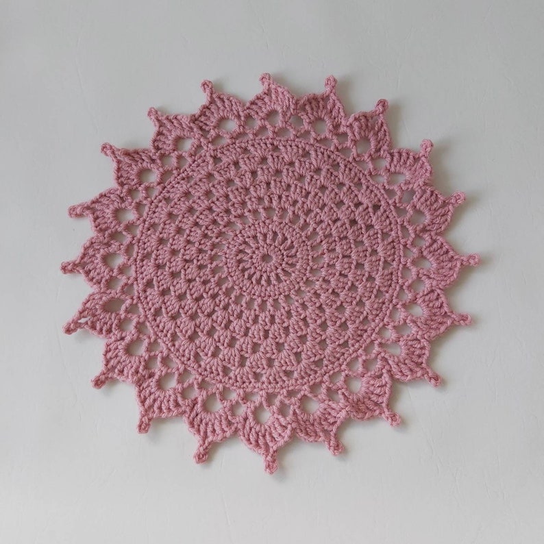 May include: A pink crocheted doily with a circular design and a scalloped edge.